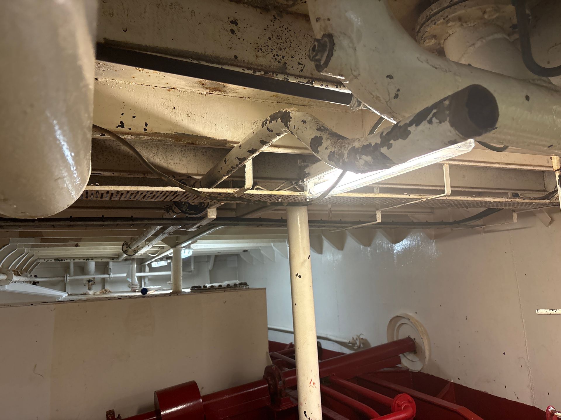 Industrial basement with white pipes, exposed ceiling, and red machinery below.