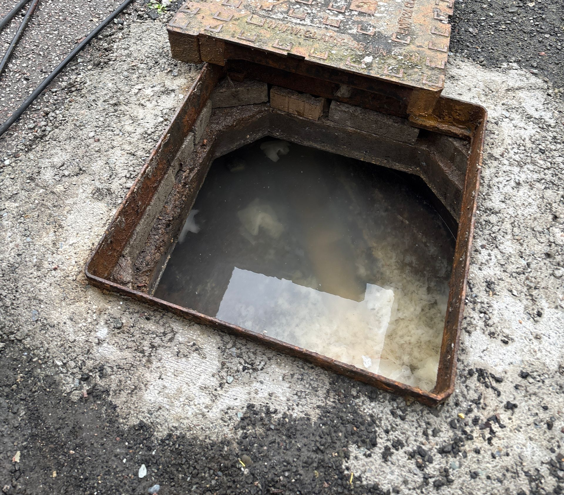Open rusty manhole cover with standing water in a concrete pavement.