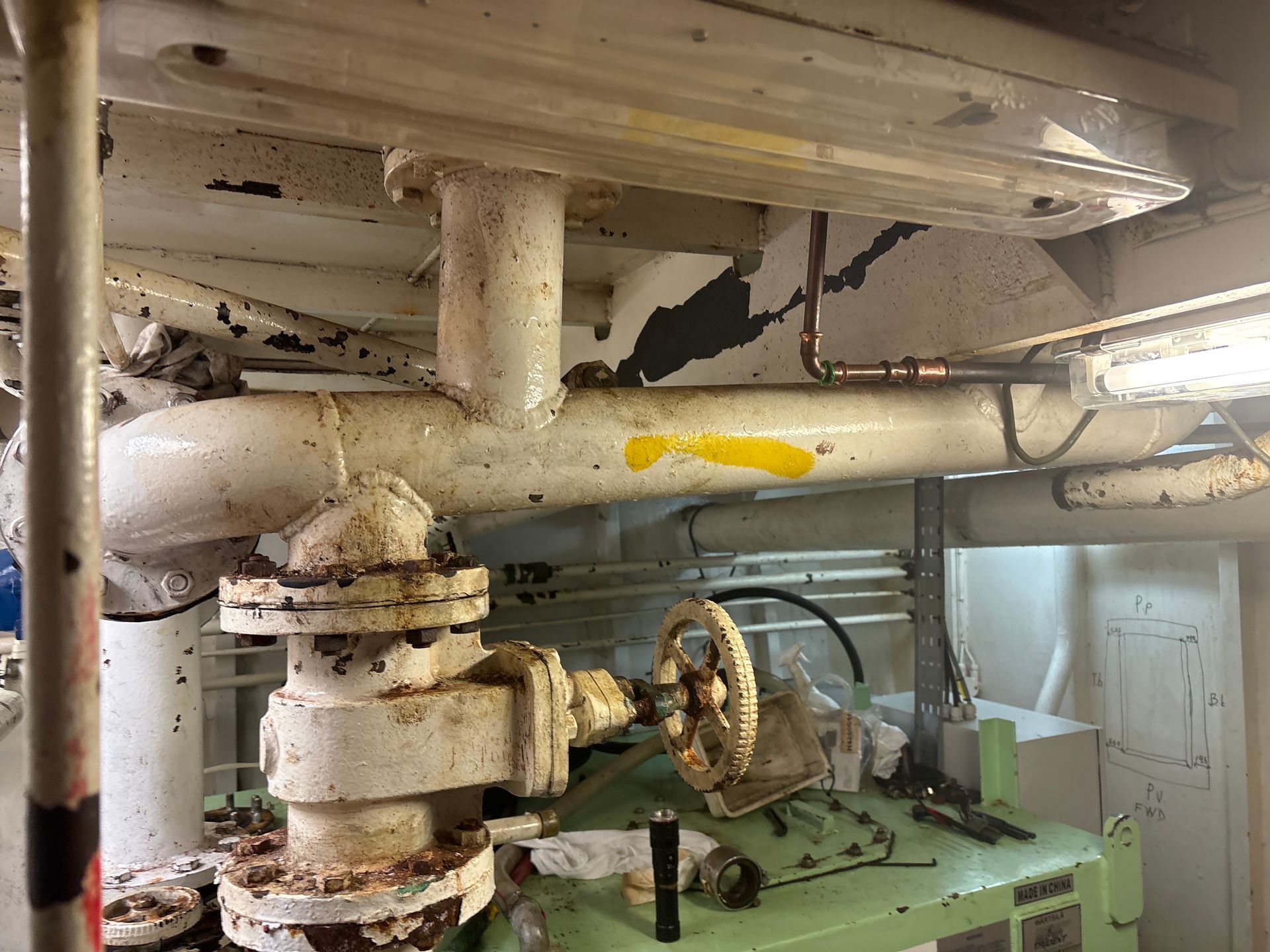 Rusty industrial pipes and a valve in a cramped basement with a green machine below.