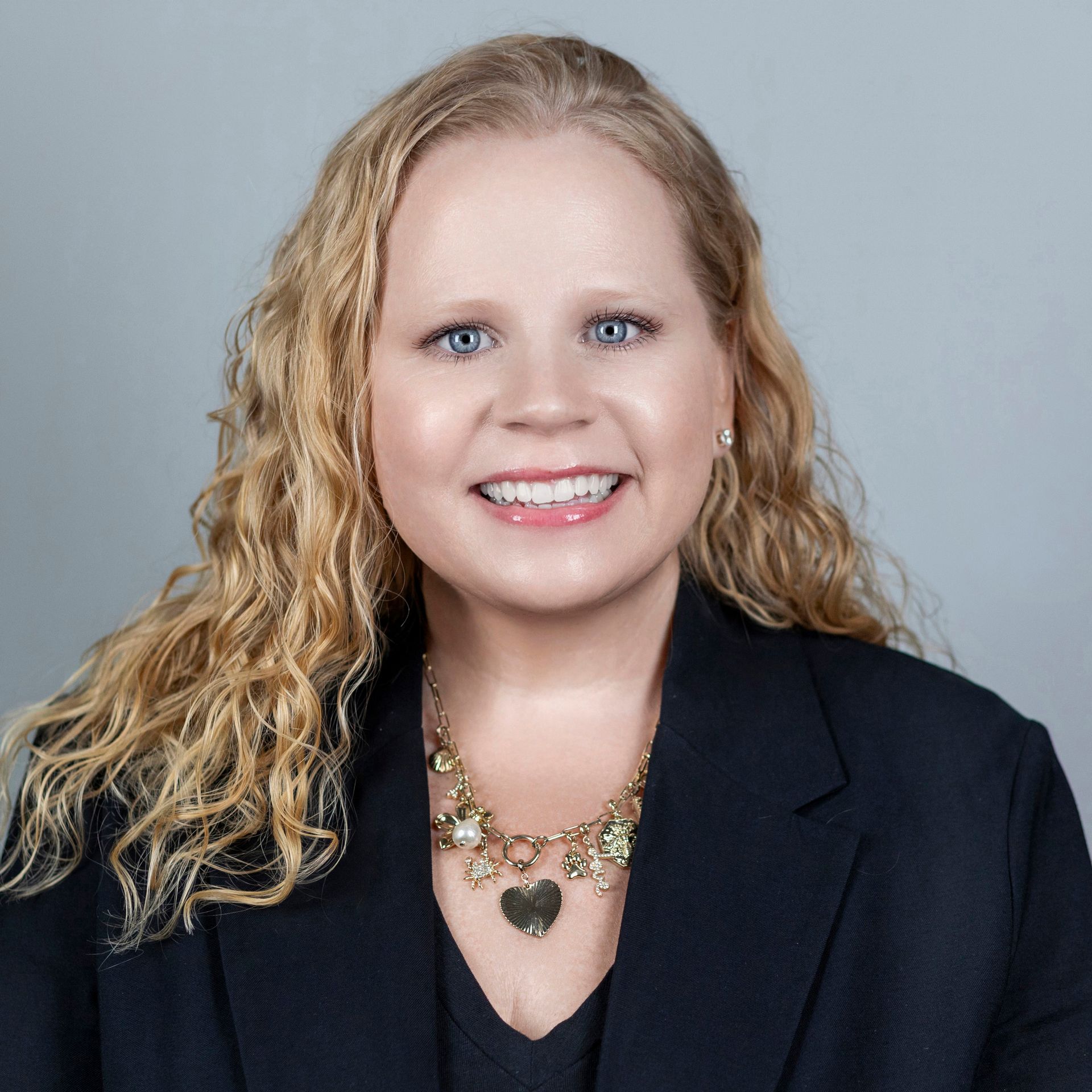 Woman with blonde curly hair smiles, wearing a black blazer and gold heart pendant necklace.