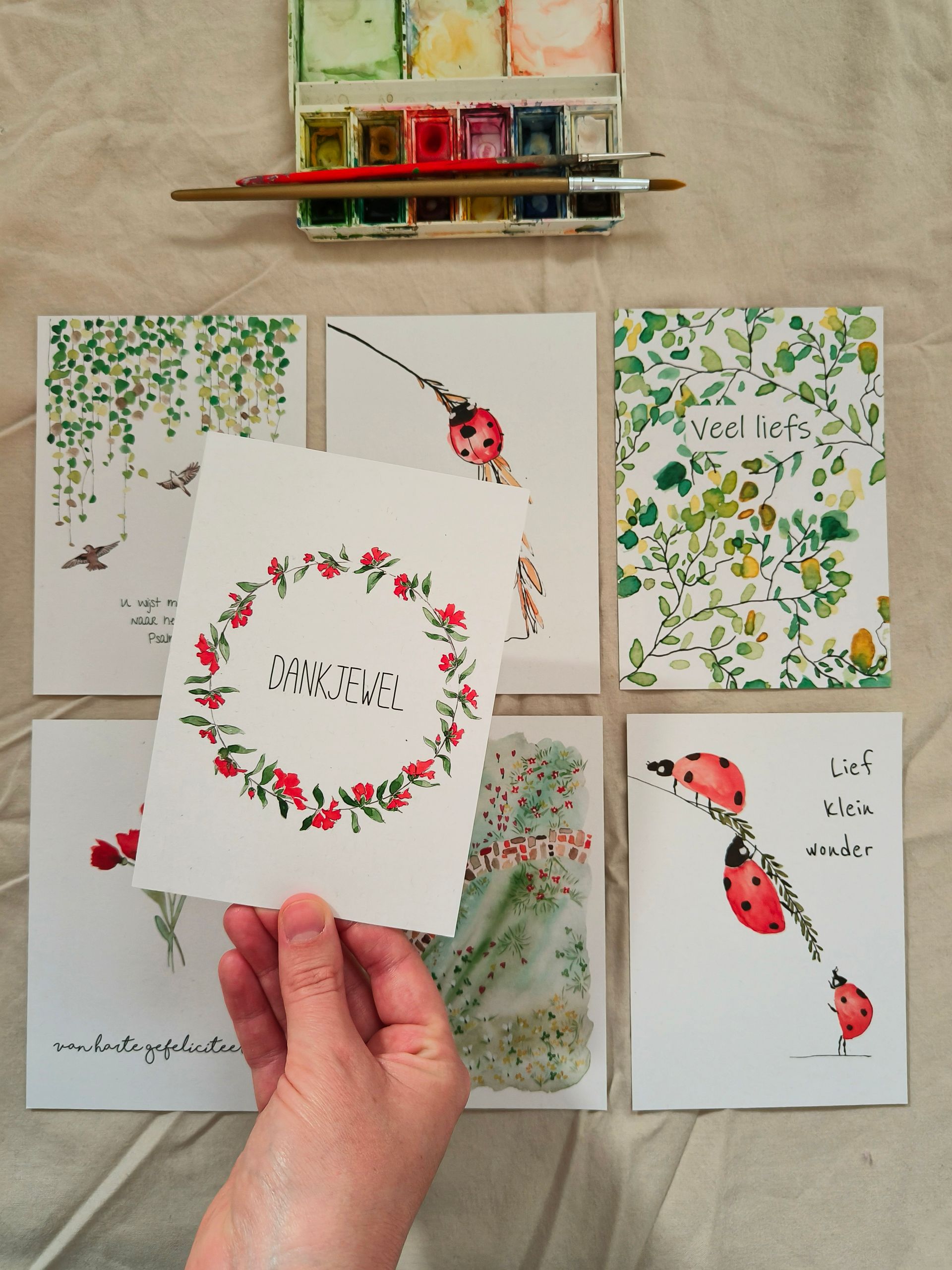 Hand die een kaartje met een bloemenkrans vasthoudt. Aquarelschilderijen van planten en lieveheersbeestjes.