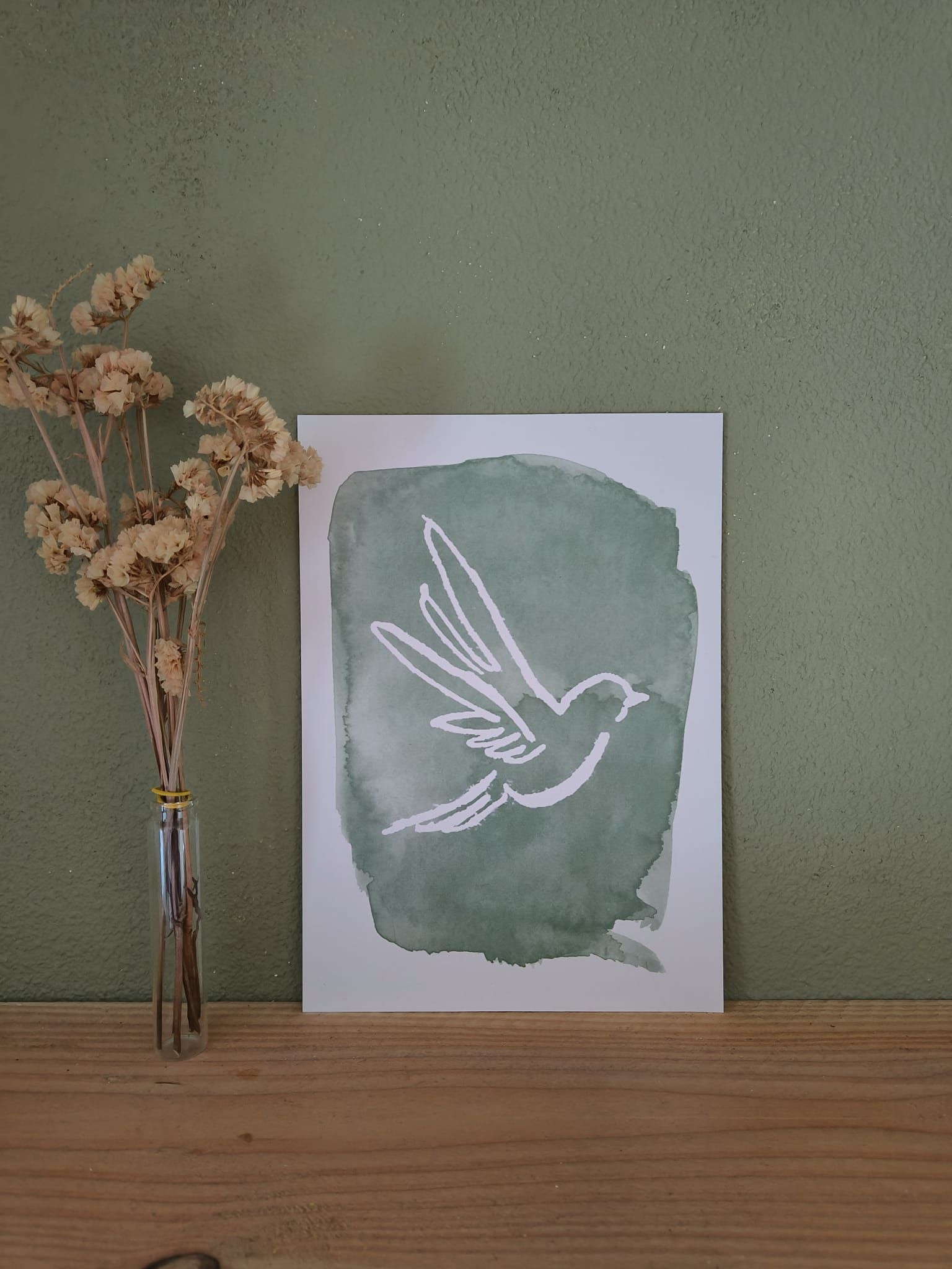 Aquarel schilderij van een witte vogel of duif in vlucht tegen een groene achtergrond. Gedroogde bloemen in een vaas ernaast.