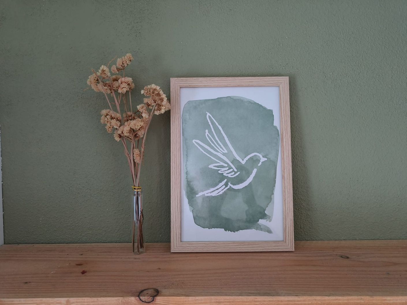 A4 poster van een vogel of duif in een houten lijst op een plank, gedroogde bloemen in een glazen vaas tegen een groene muur.