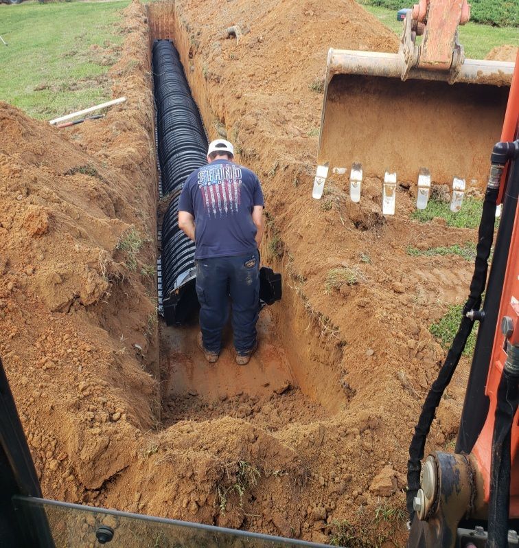 Man installing black corrugated pipe in a trench; an excavator bucket is visible in the frame.