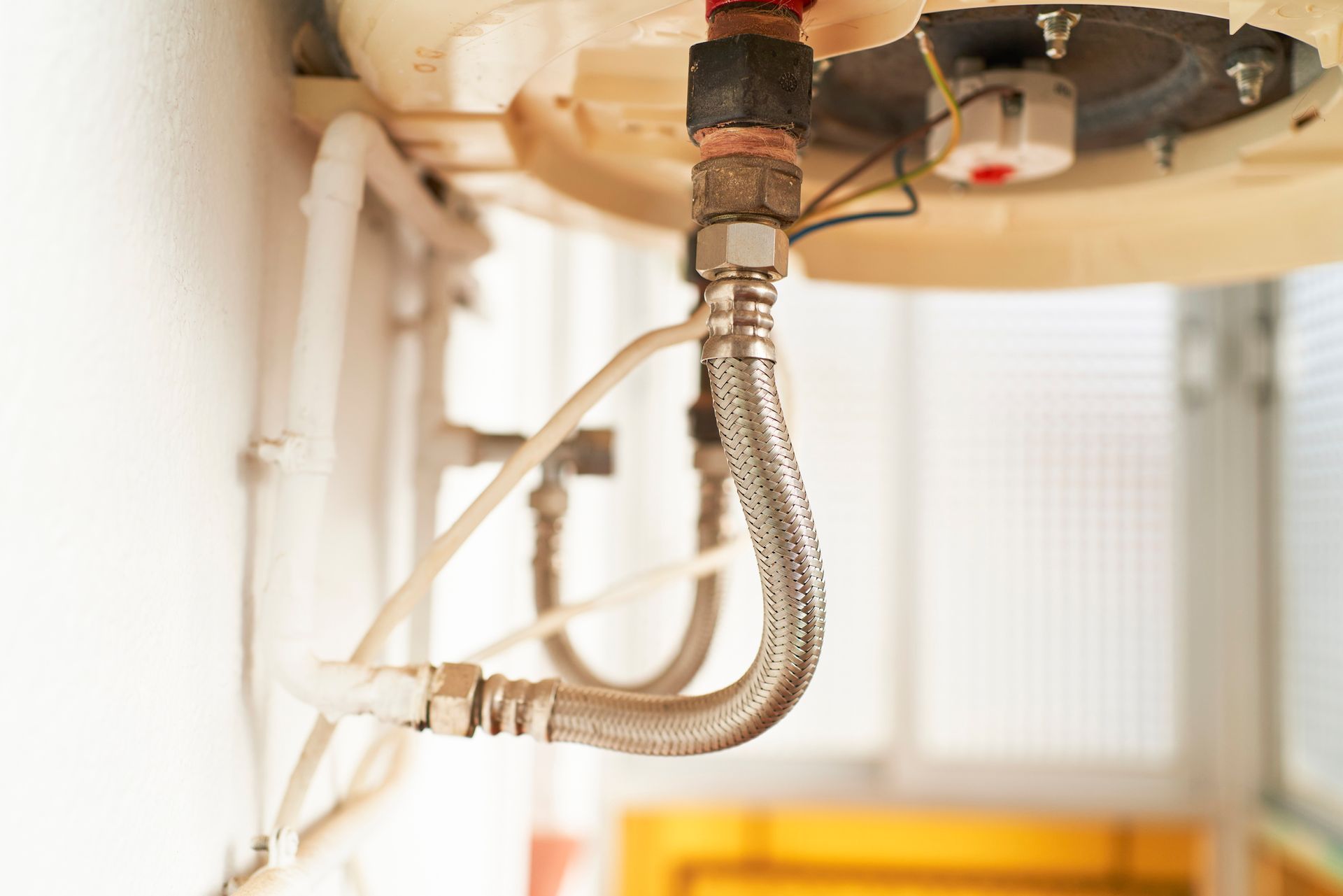 Close-up of a water heater with flexible metal supply lines and white plastic piping against a white wall.