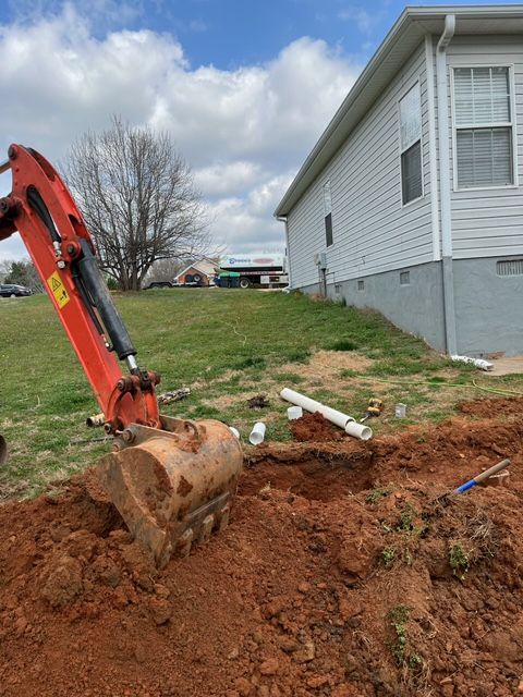 An excavator digs a trench near a house with white siding and gray foundation; orange arm, red dirt.
