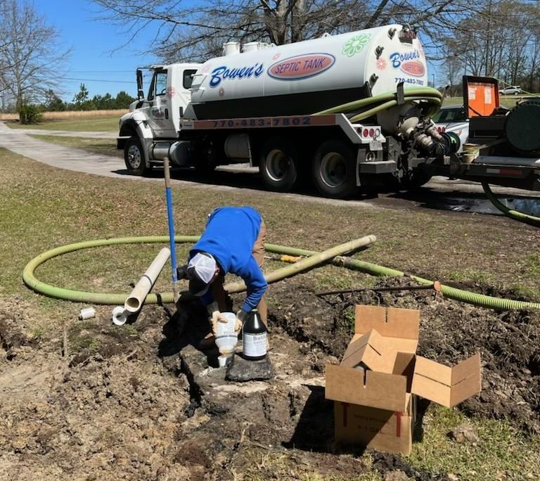 Septic tank being serviced; worker in blue, truck, pipes in dirt, sunny day.