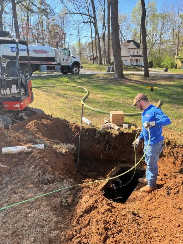 Man in blue shirt works on septic tank, excavator and truck visible.