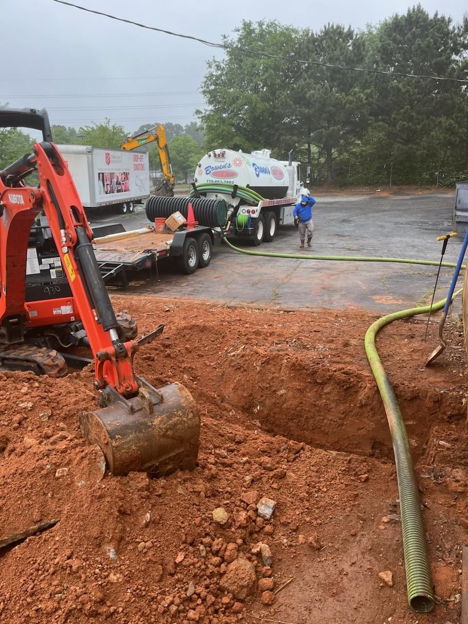 Construction site with excavator digging in red dirt, septic tank truck and worker in background.