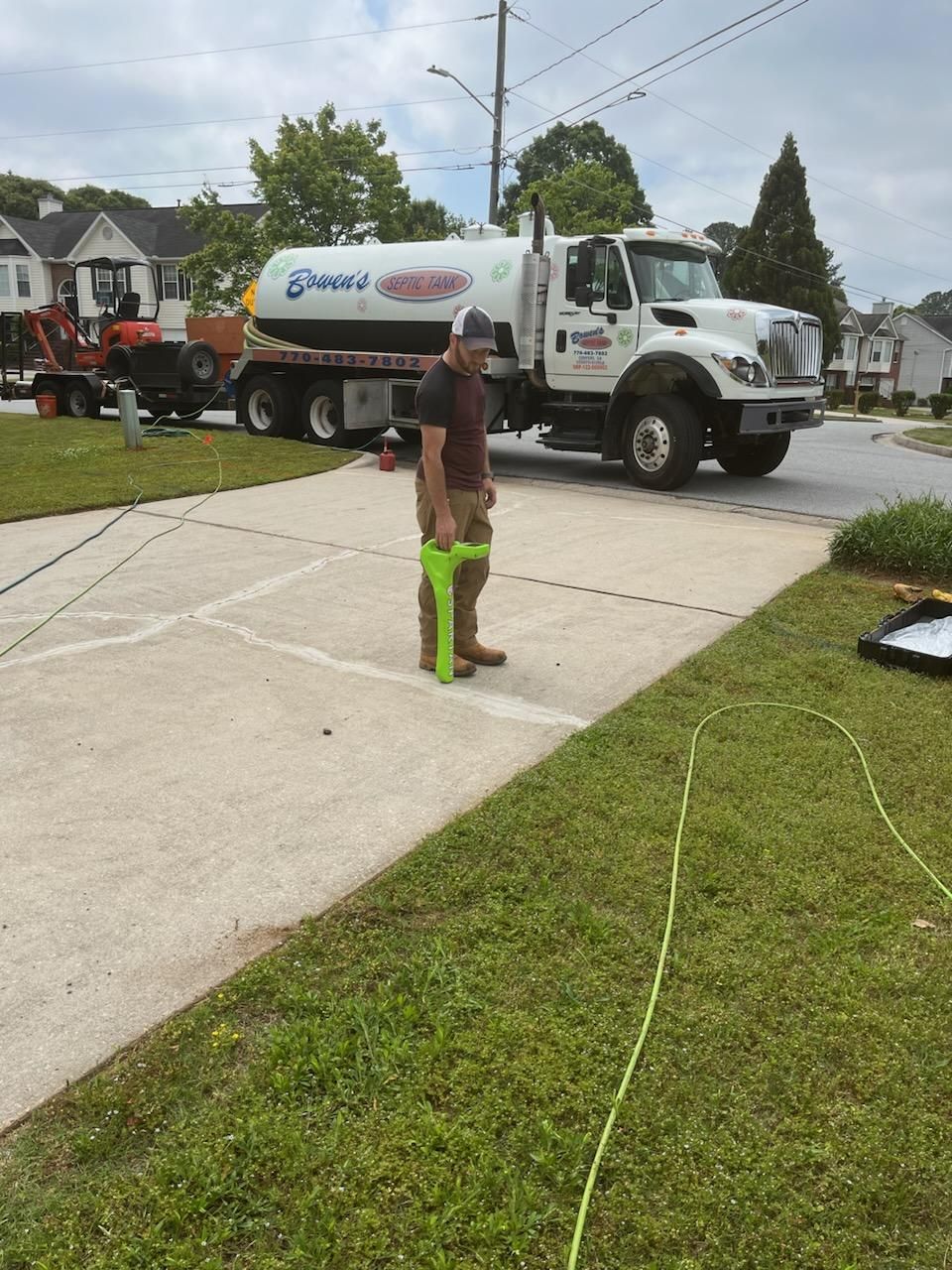 Man with a level, standing on a driveway with a septic truck parked nearby. Green grass and houses in the background.