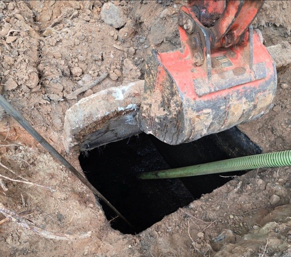 An excavator bucket unearths a dark pit, with a green hose extended into it. An excavator bucket unearths a dark pit, with a green hose extended into it.