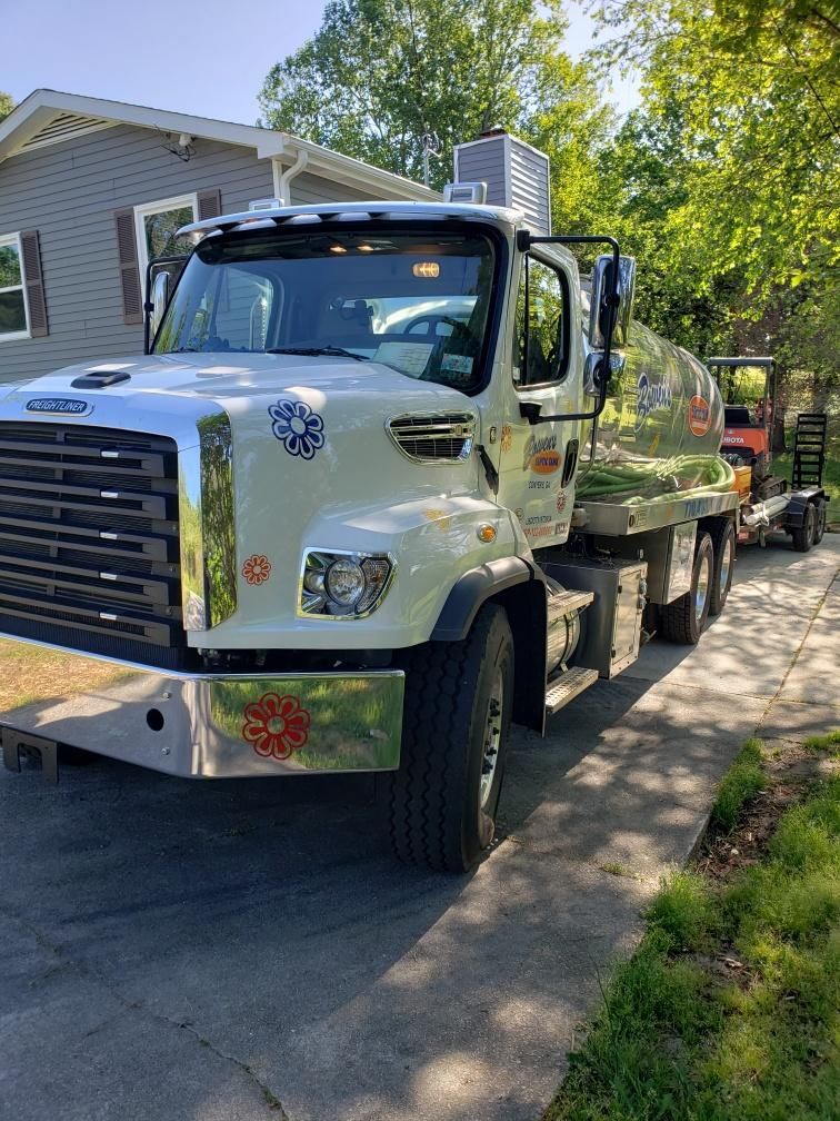 White tanker truck with a trailer, parked on a driveway near a house and trees.