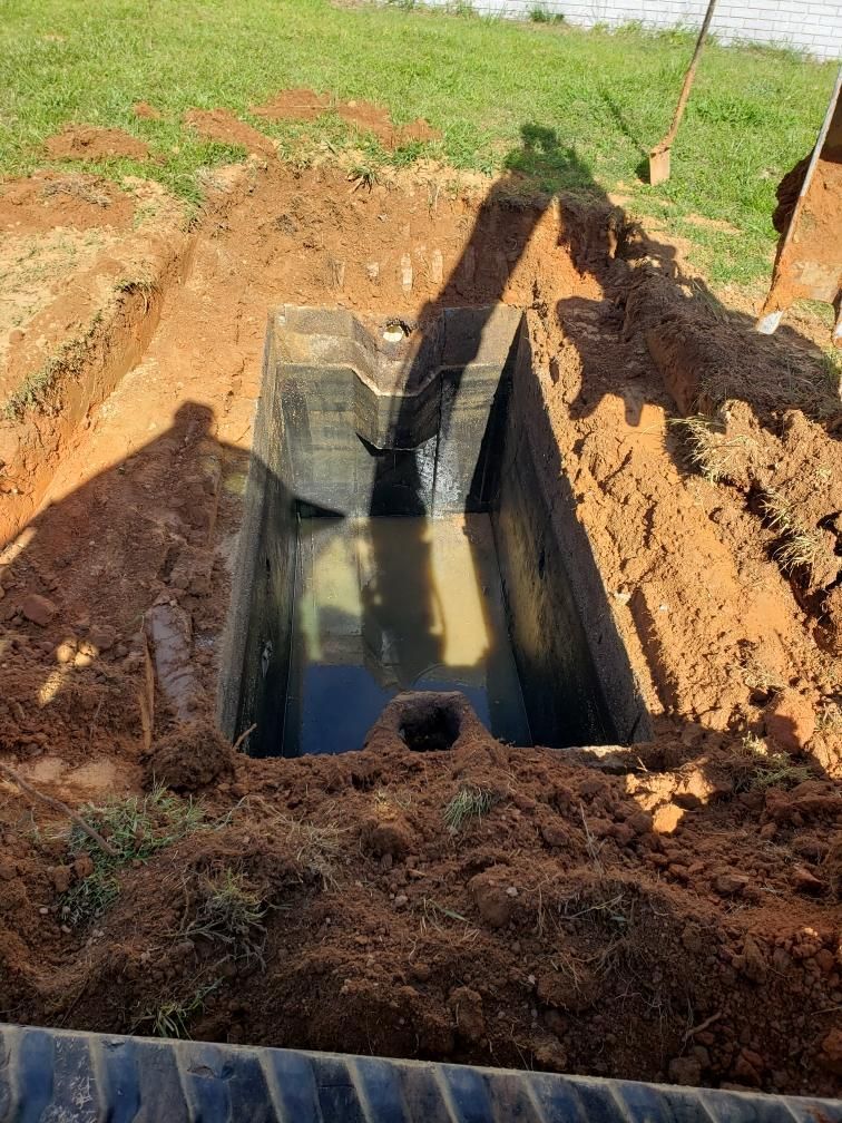 A rectangular concrete tank sits in a dirt excavation, filled with water. Green grass and soil surround it.