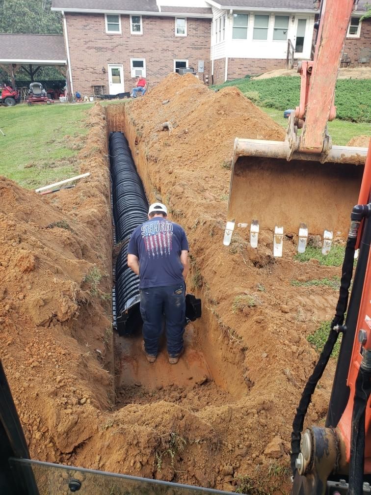 Man in a trench installing black drainage pipe near a house with an excavator.