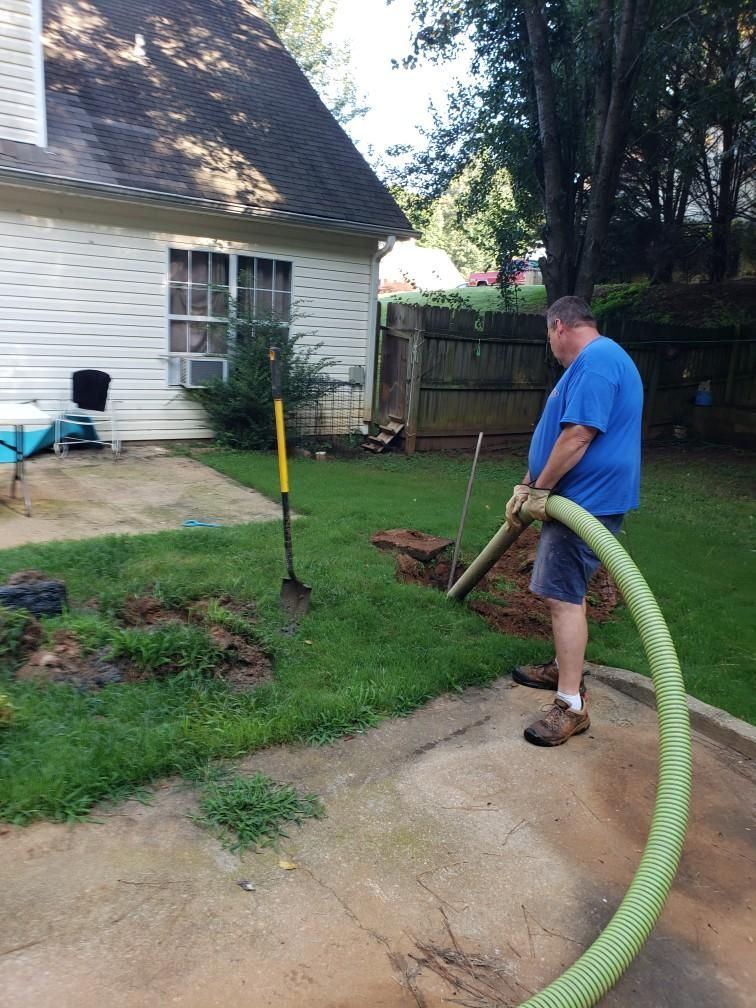 Man in blue shirt and shorts using a large green hose in a backyard. Shovel nearby.