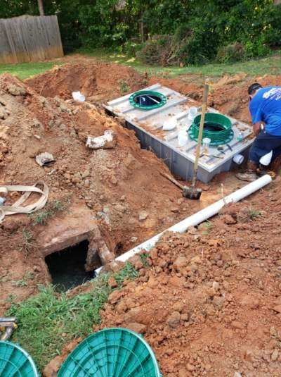 A septic system being installed in a backyard. A worker connects pipes to a buried tank in a dirt pit.