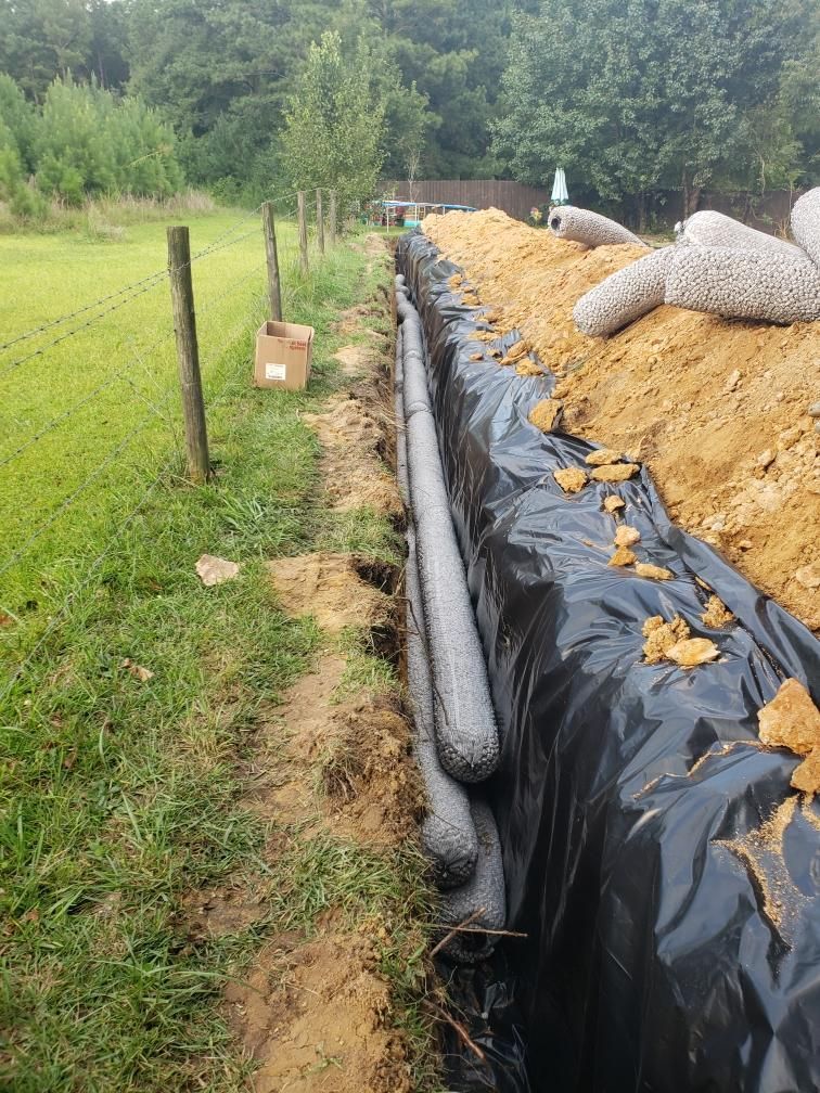 A trench with drainage tubes wrapped in mesh, lined with black fabric, alongside a grassy area, fence, and soil.