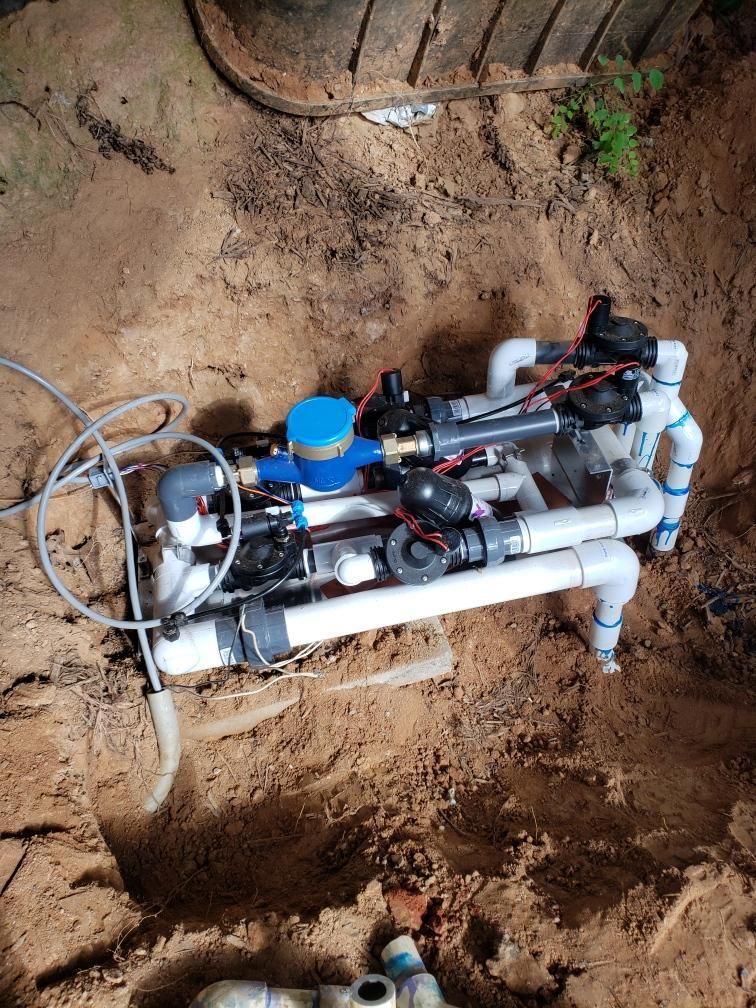 Irrigation system with PVC pipes, valves, and electrical components, installed in a sandy pit.