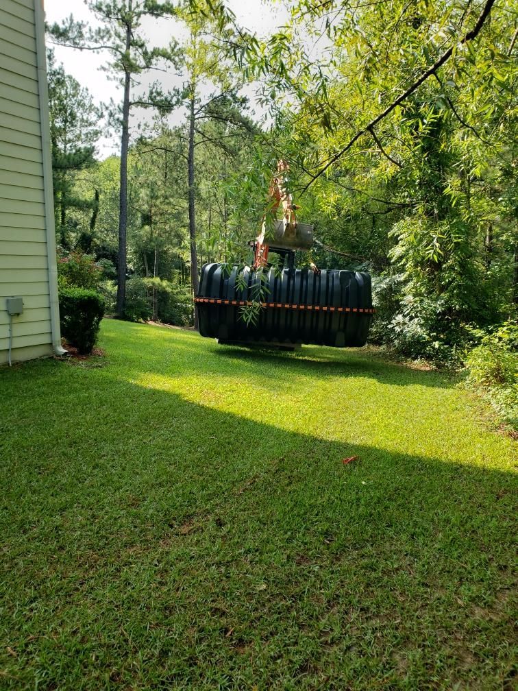 Septic tank lifted by machinery above a grassy yard near trees and a house.