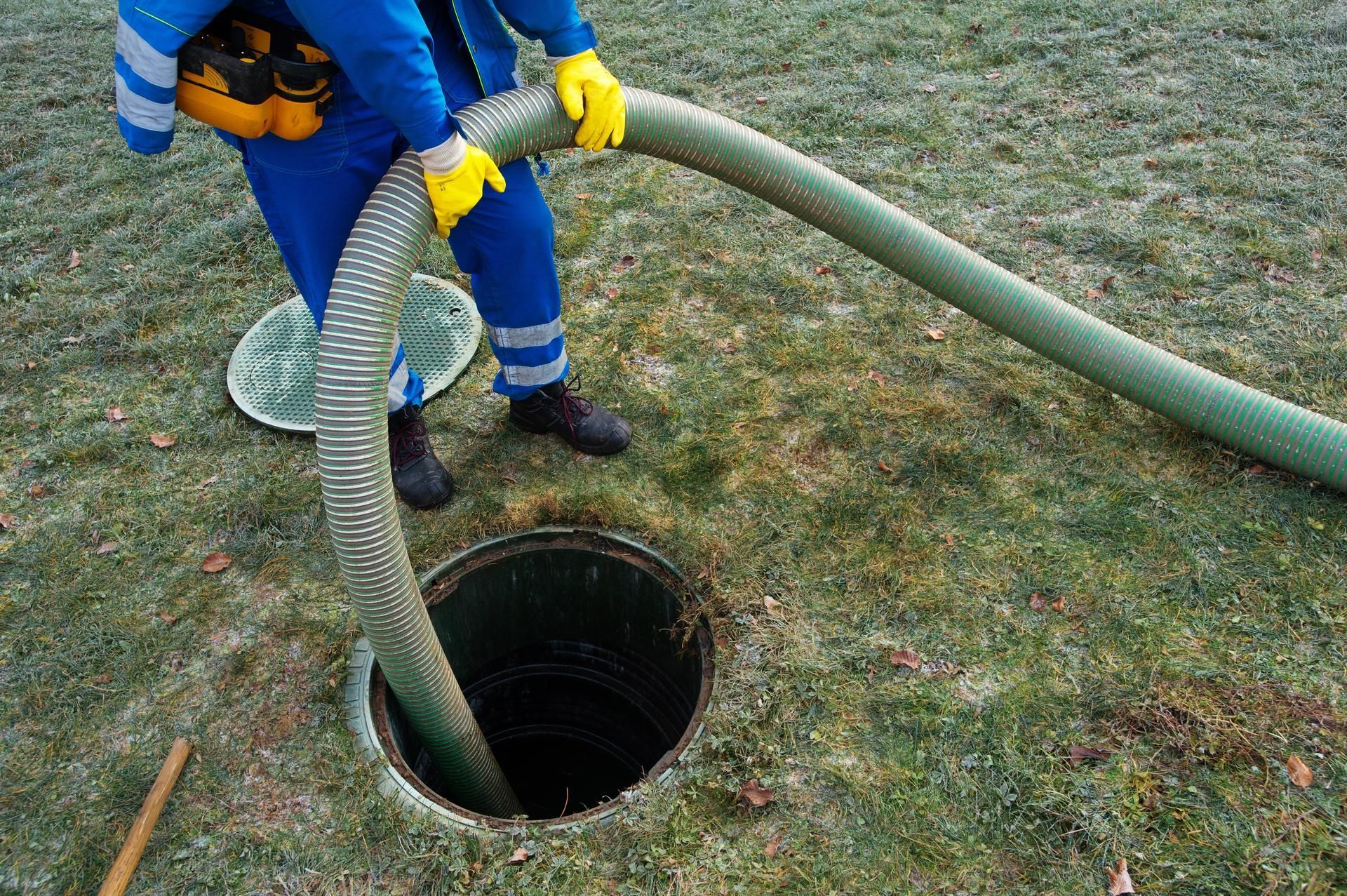 Person in blue overalls with a hose at a septic tank opening, on green grass.