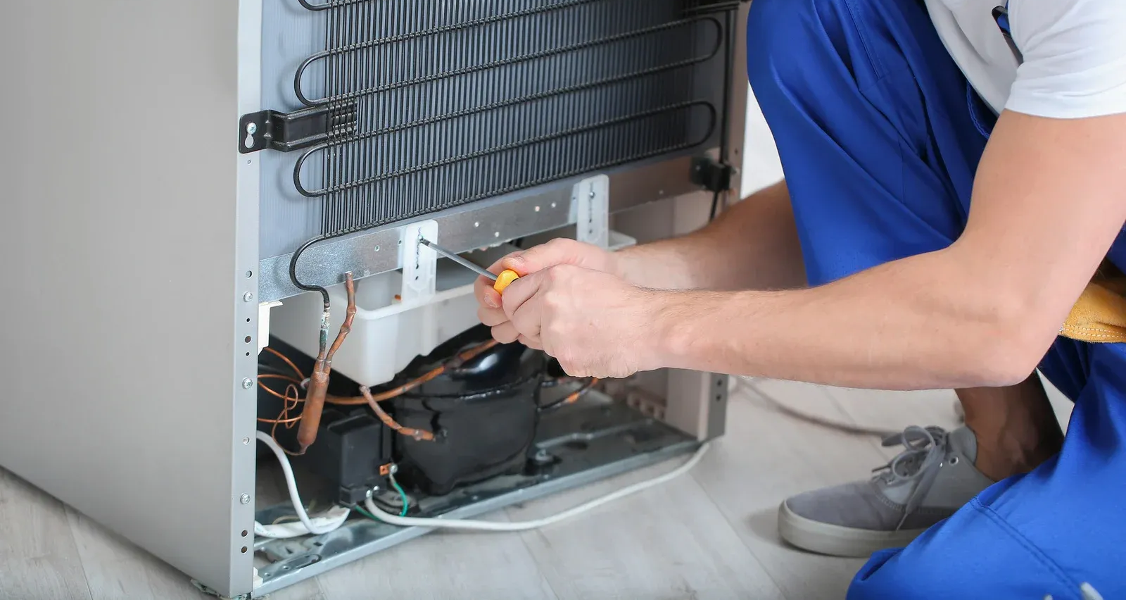 Person in blue overalls repairs a refrigerator with a screwdriver.