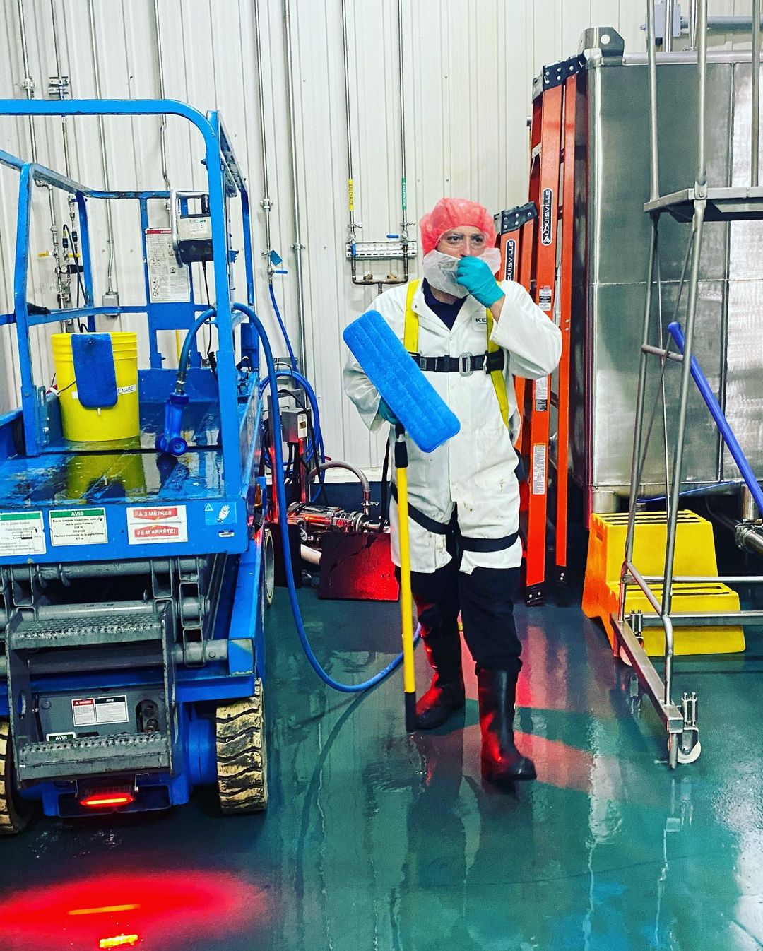 Une personne en tenue de protection tient une serpillière dans un environnement industriel, près d'un pont élévateur