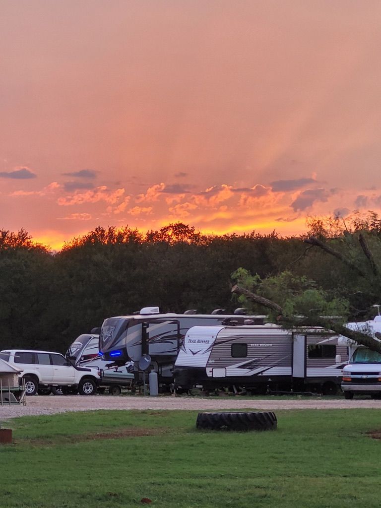Campground at sunset with RVs, green grass, and trees. Sky is orange and yellow.