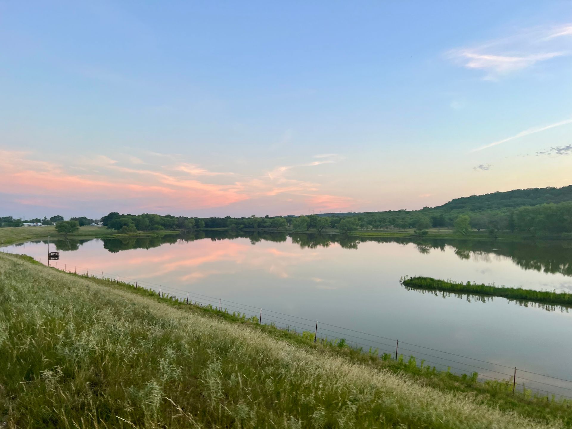 A large body of water surrounded by grass and trees with a sunset in the background.