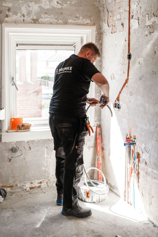 Een arbeider boort in een muur tijdens een huisrenovatie; in een gestripte ruimte zijn de bedrading en gereedschappen zichtbaar.