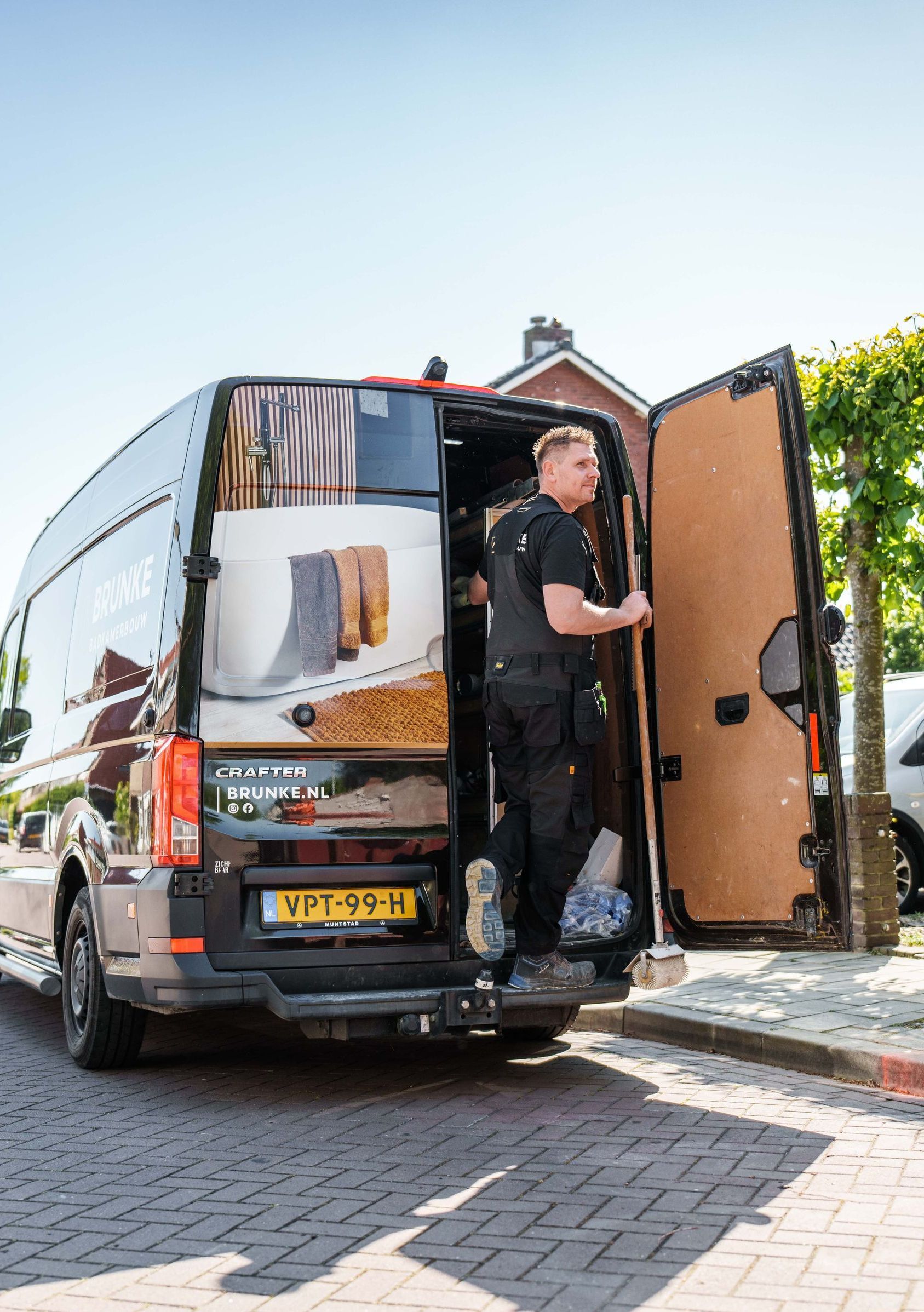Een bezorger laadt dozen uit een zwarte bestelbus die geparkeerd staat op een straat met kinderkopjes.