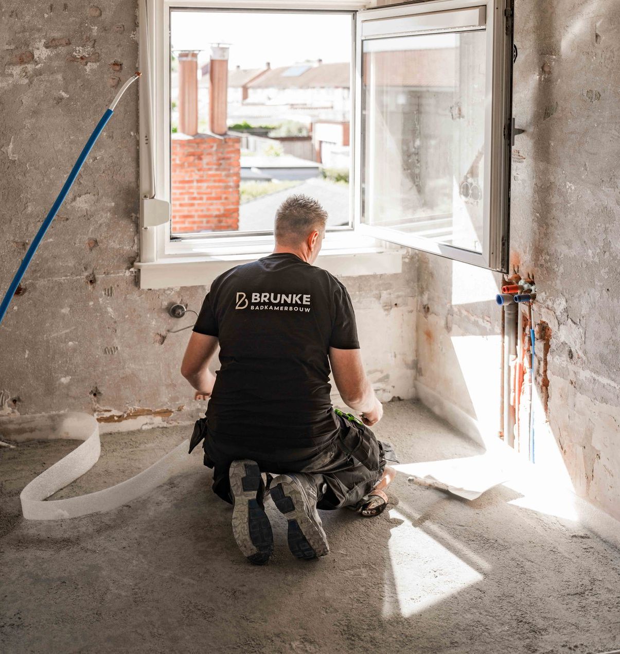 Een arbeider knielt in een stoffige ruimte tijdens een renovatie, met zijn gezicht naar een open raam waar het zonlicht doorheen stroomt.