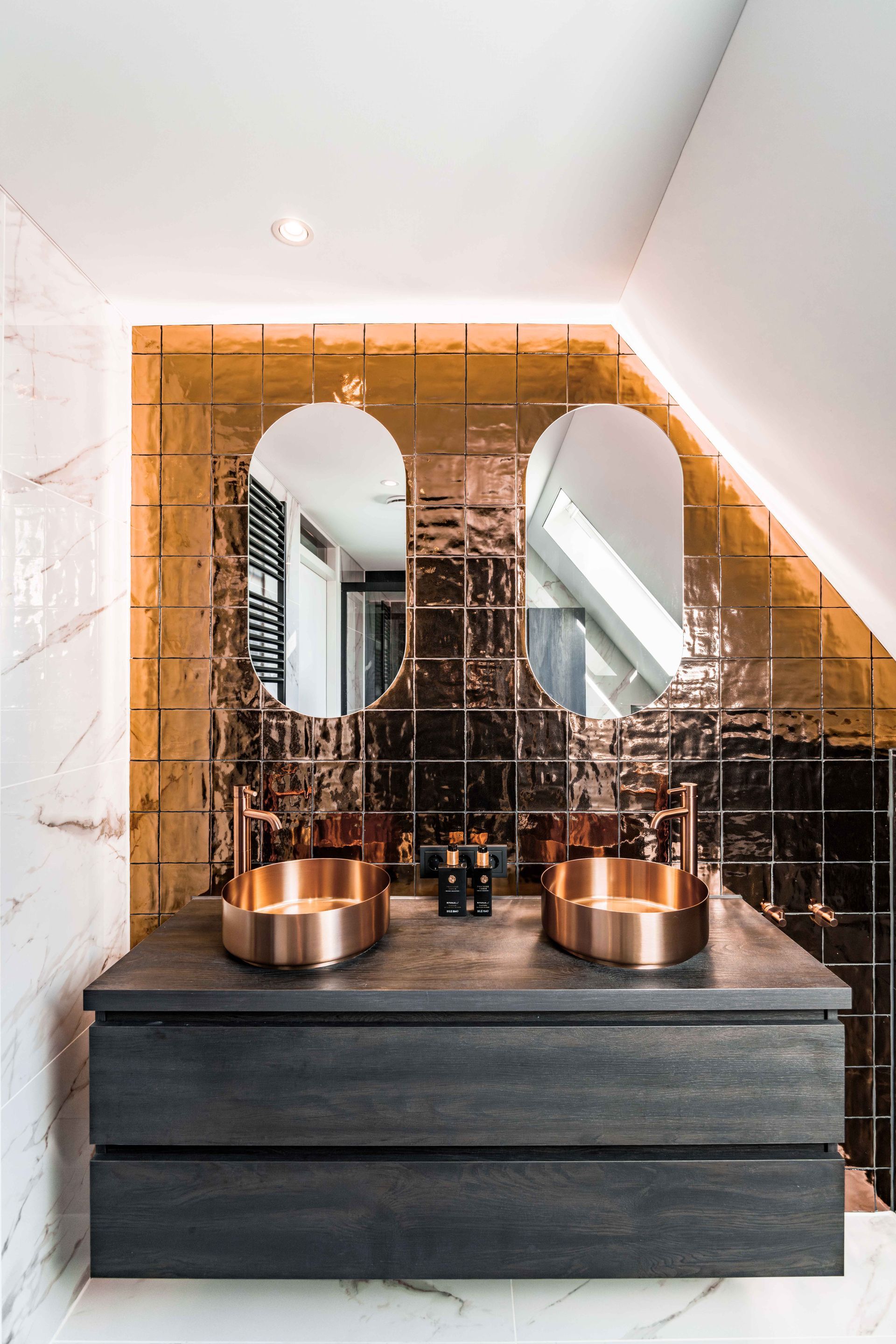 Moderne badkamermeubel met koperen wasbakken, een gebogen spiegel en warme ledverlichting in een stijlvolle badkamer.