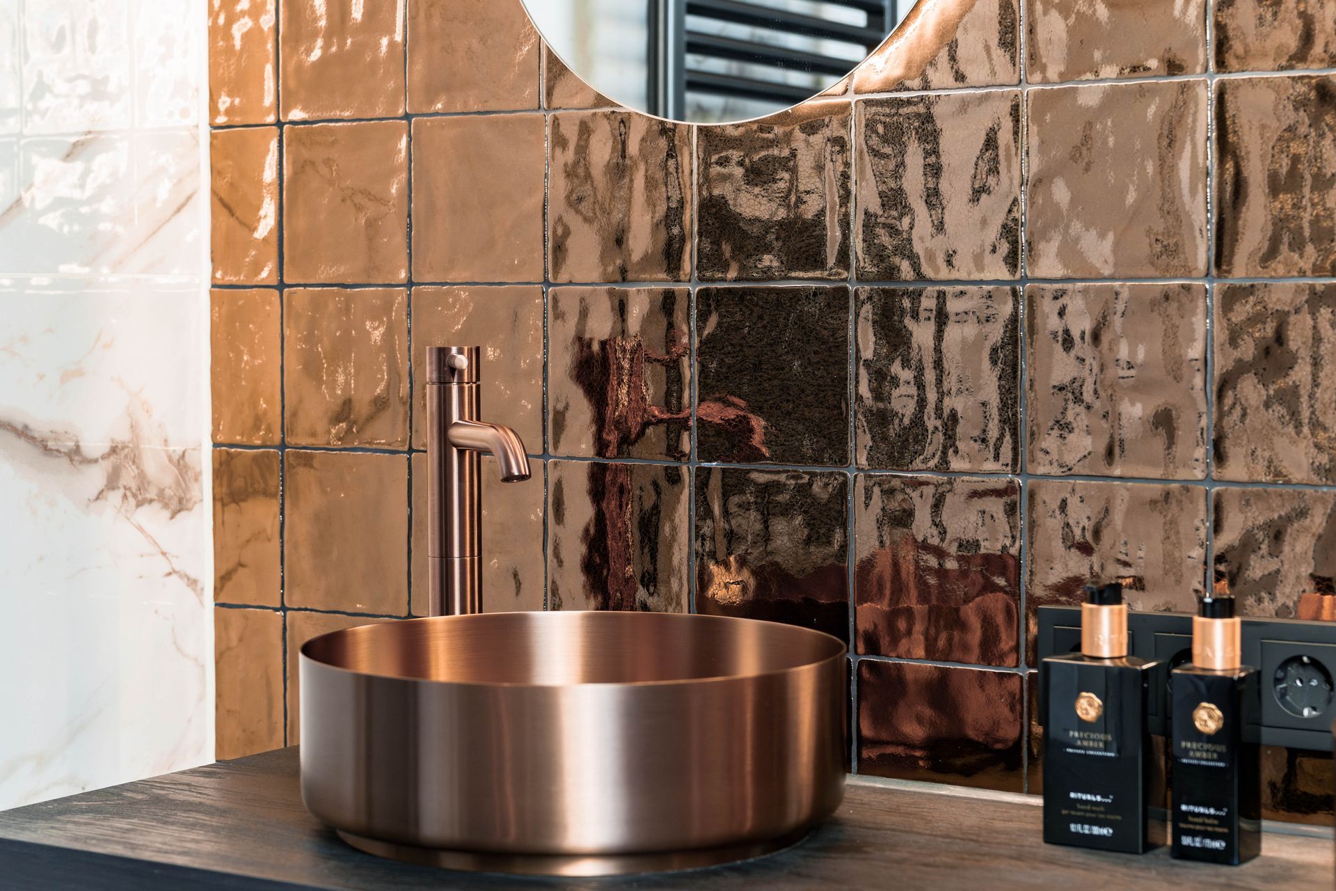 Koperen waskom op een marmeren aanrechtblad met chromen kraan en donkere toiletartikelen in een moderne badkamer.