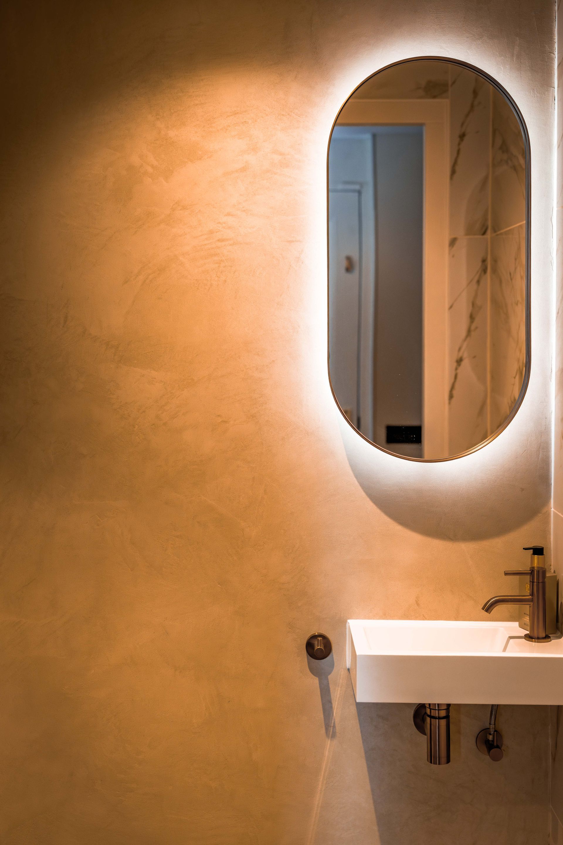Badkamerwastafel en ovale spiegel tegen een beige muur met warme verlichting.