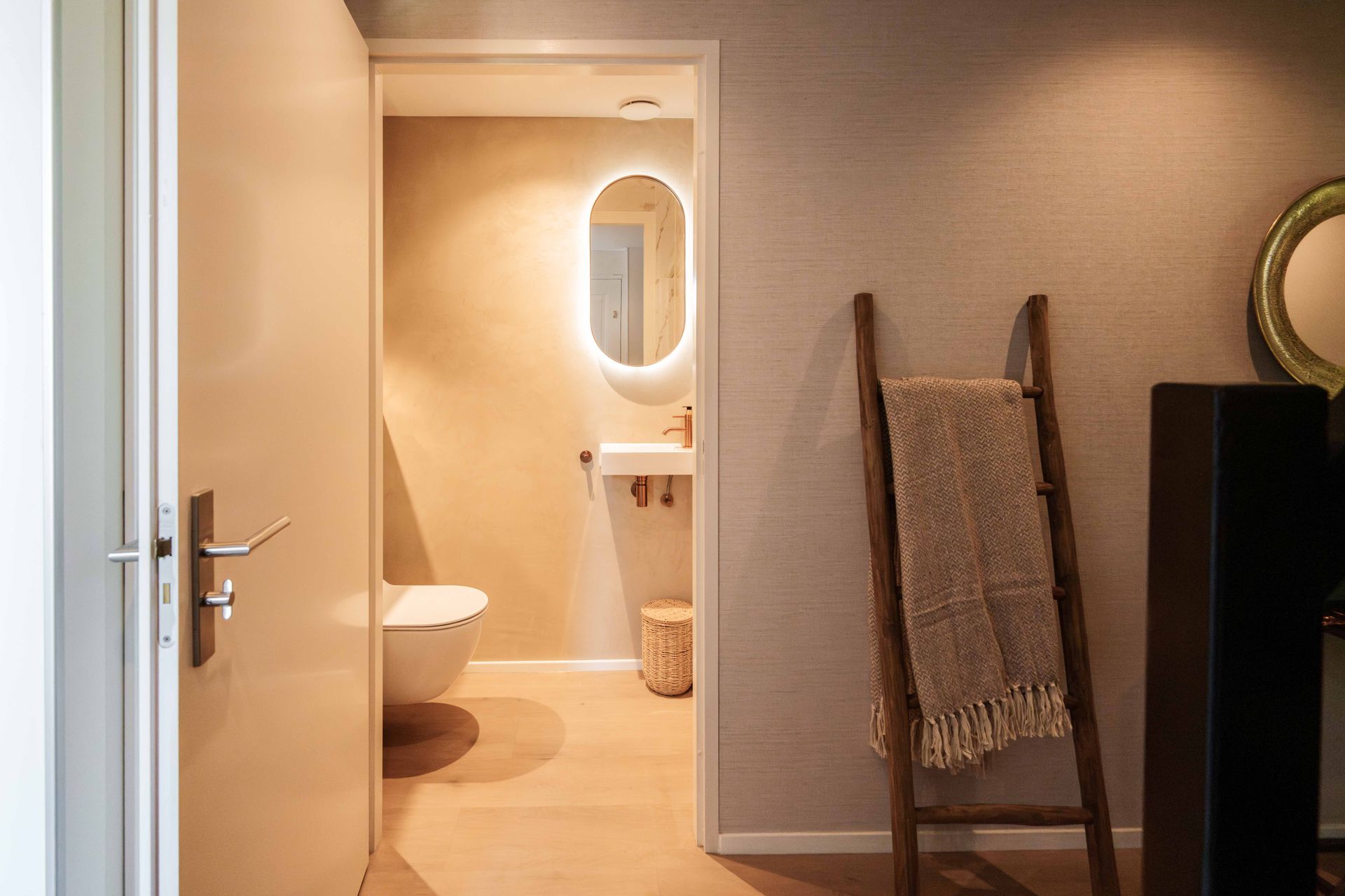 Open badkamerdeur met toilet, ovale spiegel en warme verlichting in een beige gang.