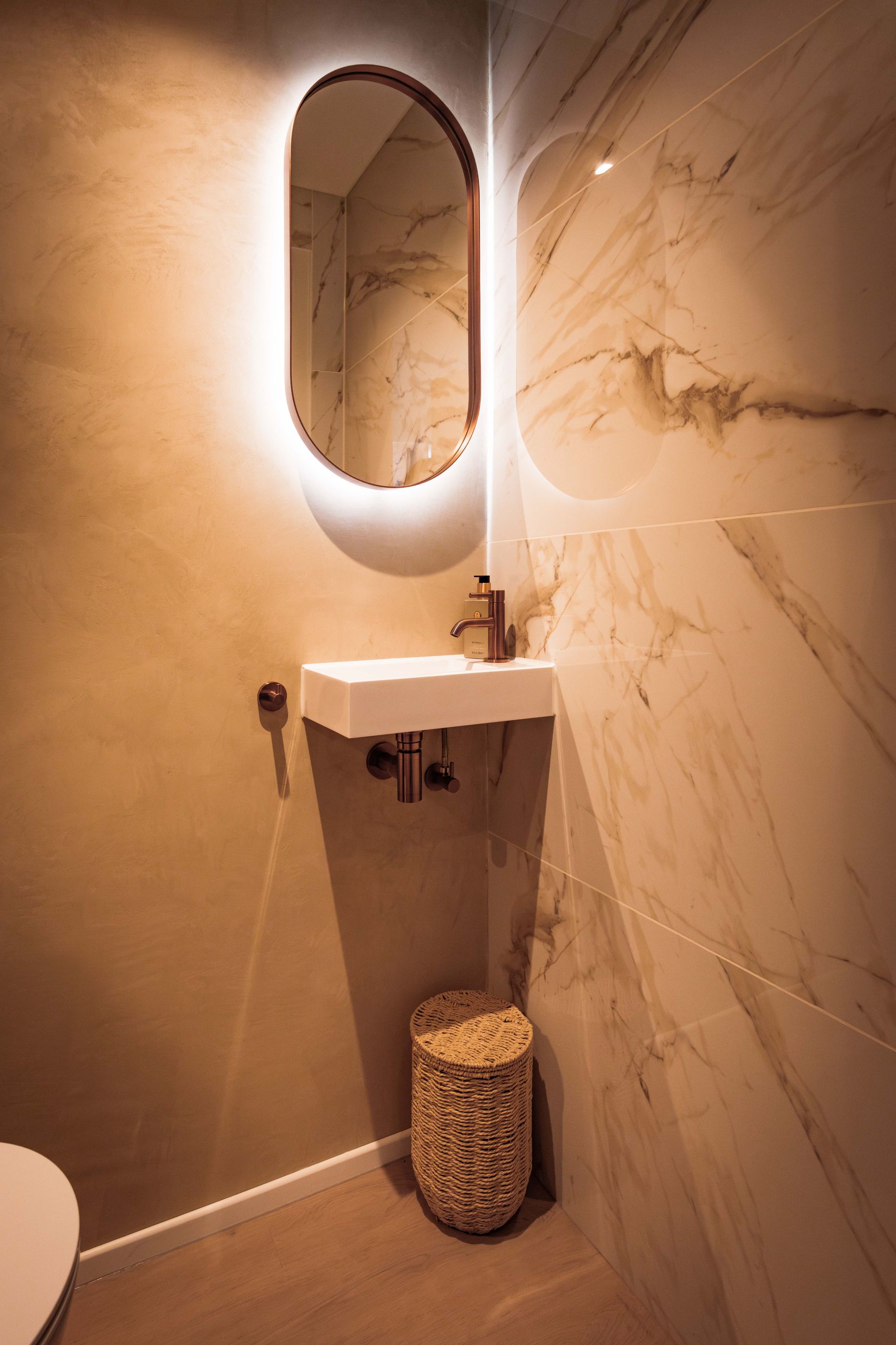 Moderne badkamerkast met ovale spiegel met achtergrondverlichting, witte wastafel, italiaans sierpleister en rieten afvalbak.