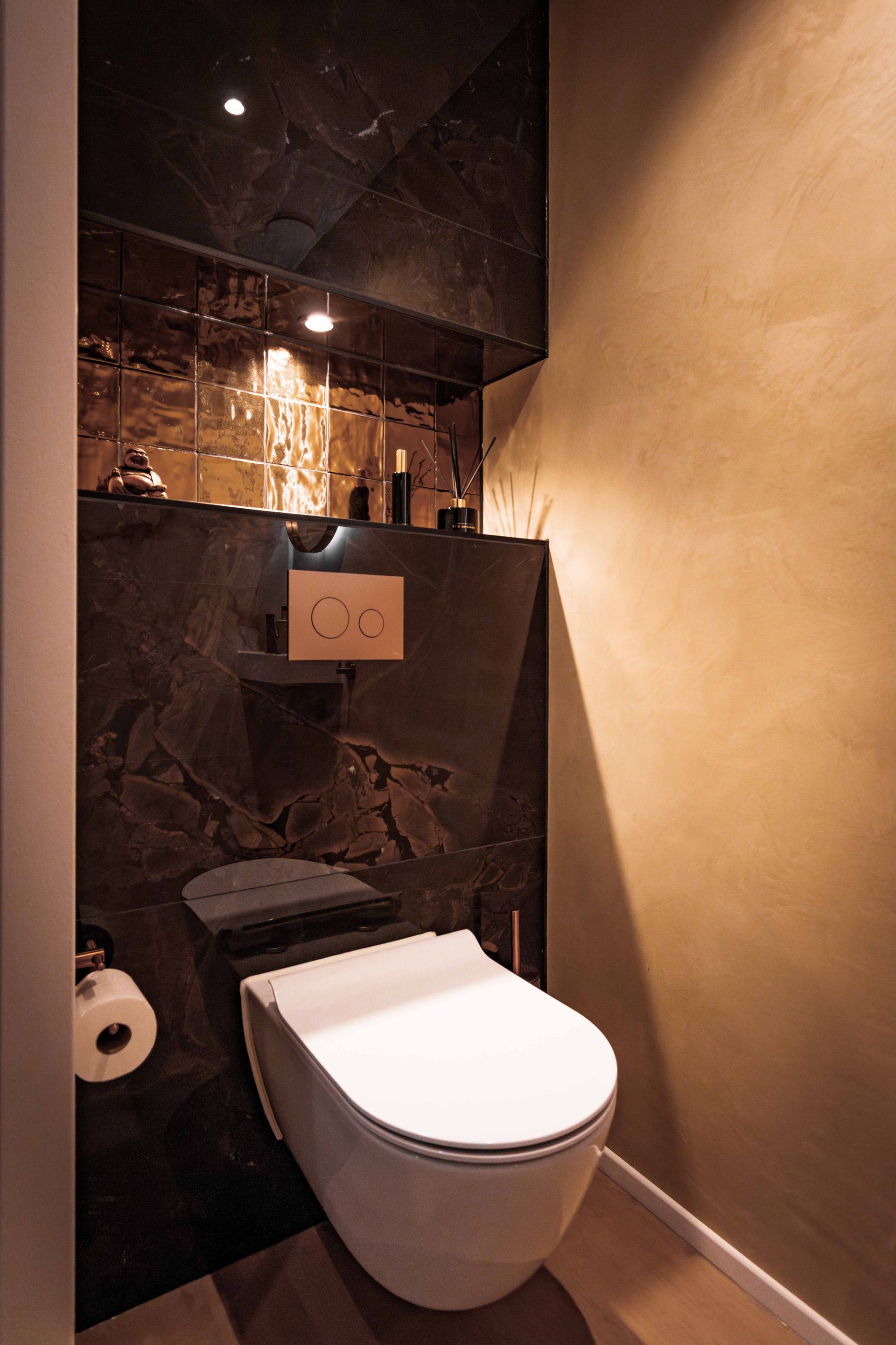 Moderne badkamer met een wit wandtoilet en donkere marmeren muren, verlicht door warme plafondlampen.