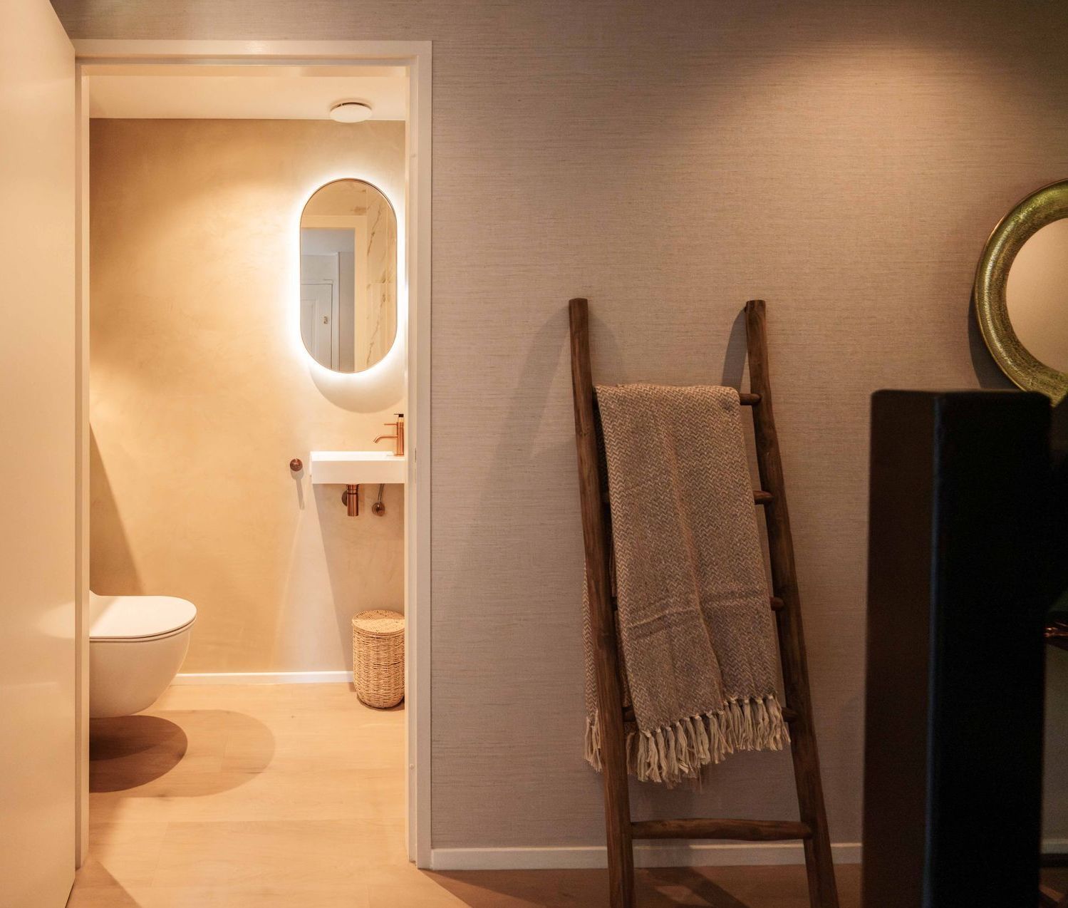 Badkamerdeur in warme tinten met ovale spiegel, wastafel, toilet en een ladder tegen de muur.