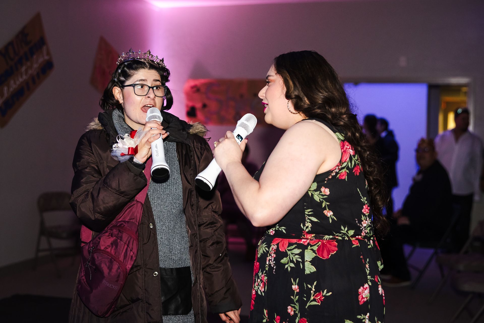 Two people sing into microphones at a party: one in black and burgundy, the other in a floral dress.