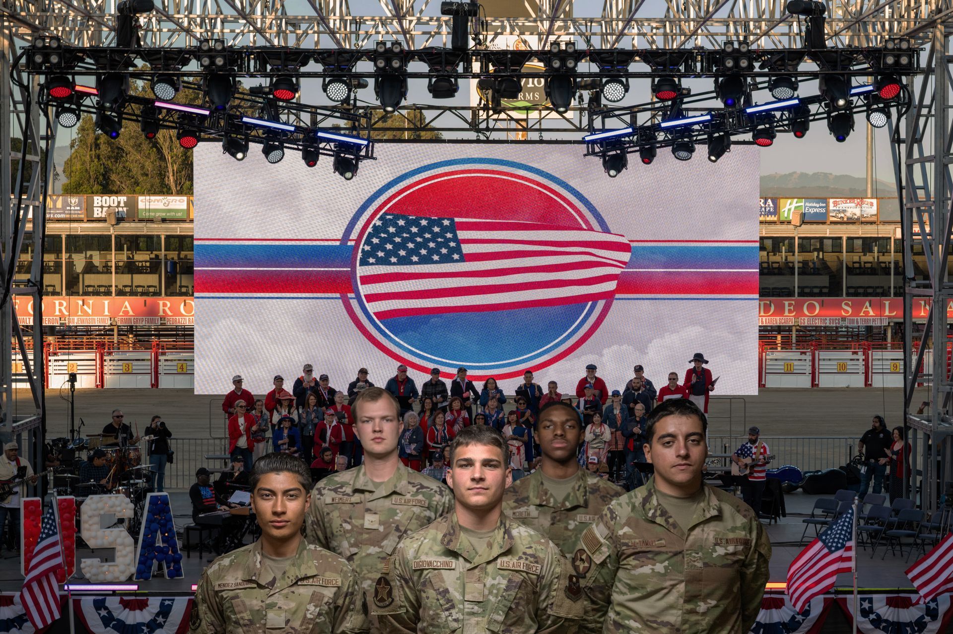 Uniformed soldiers on stage in front of a large American flag display at an outdoor event