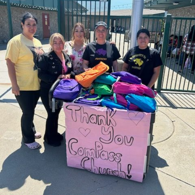 People standing behind a pink thank-you sign with colorful backpacks in front of a fenced entrance.