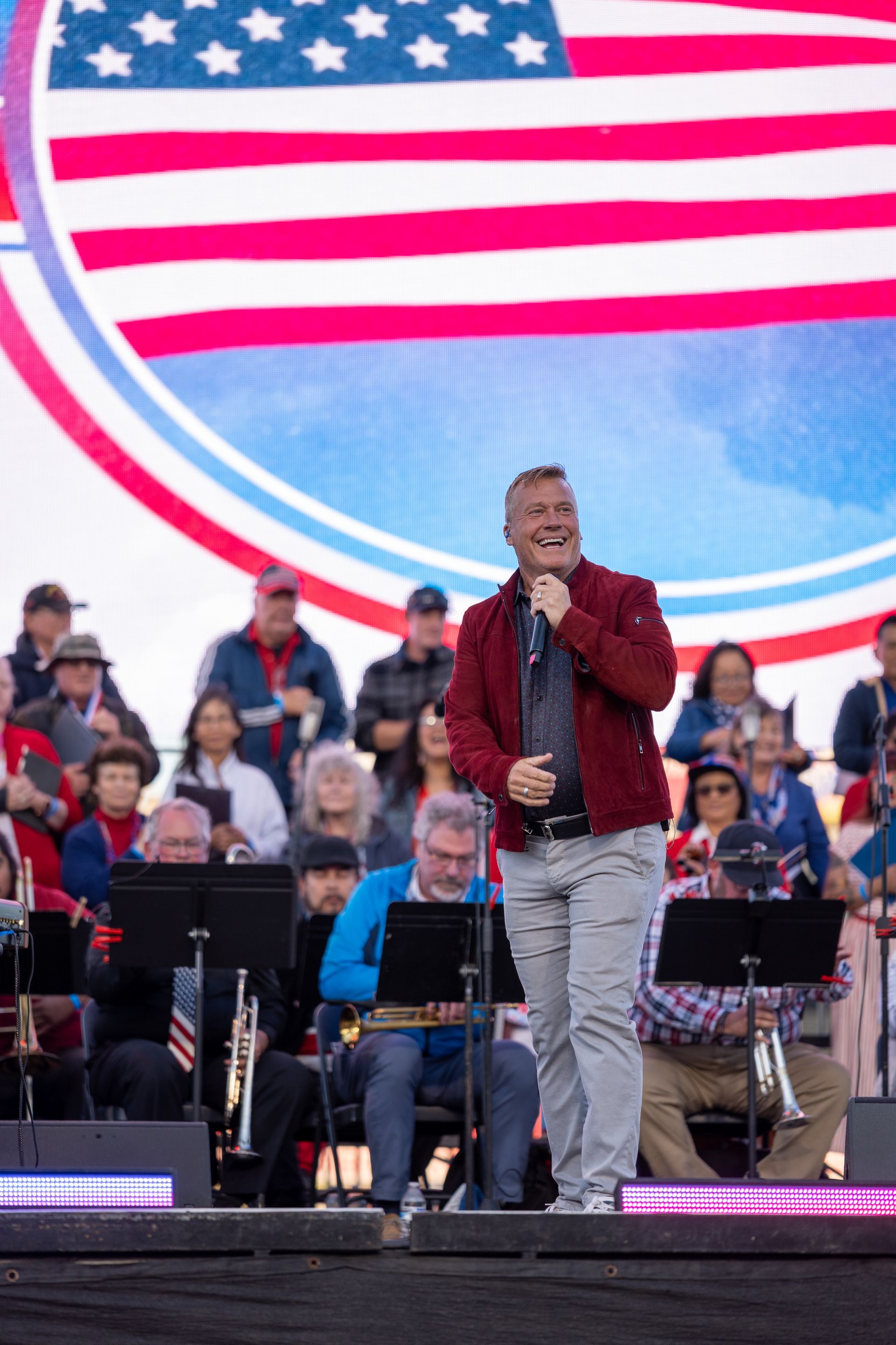 Man singing on stage, red jacket, gray pants. American flag backdrop. Band and crowd.