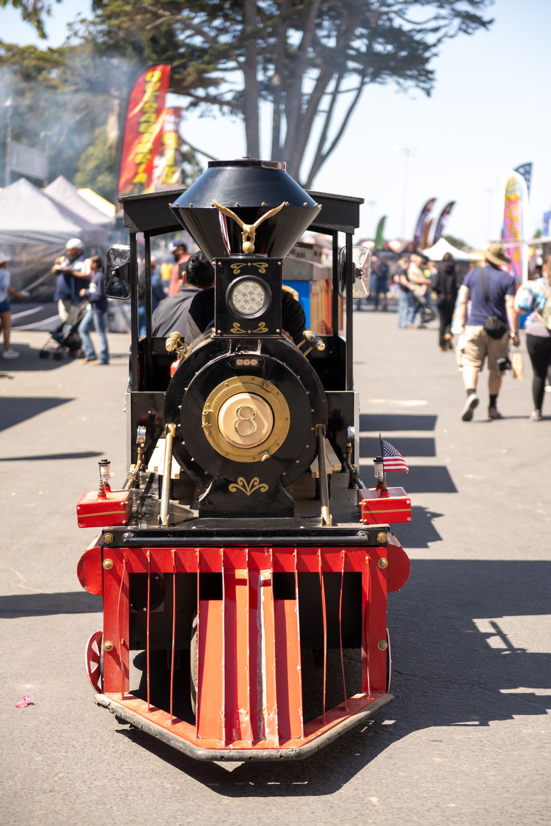 Miniature black and red steam train on a paved road, outdoor fair setting. Smoke rising.