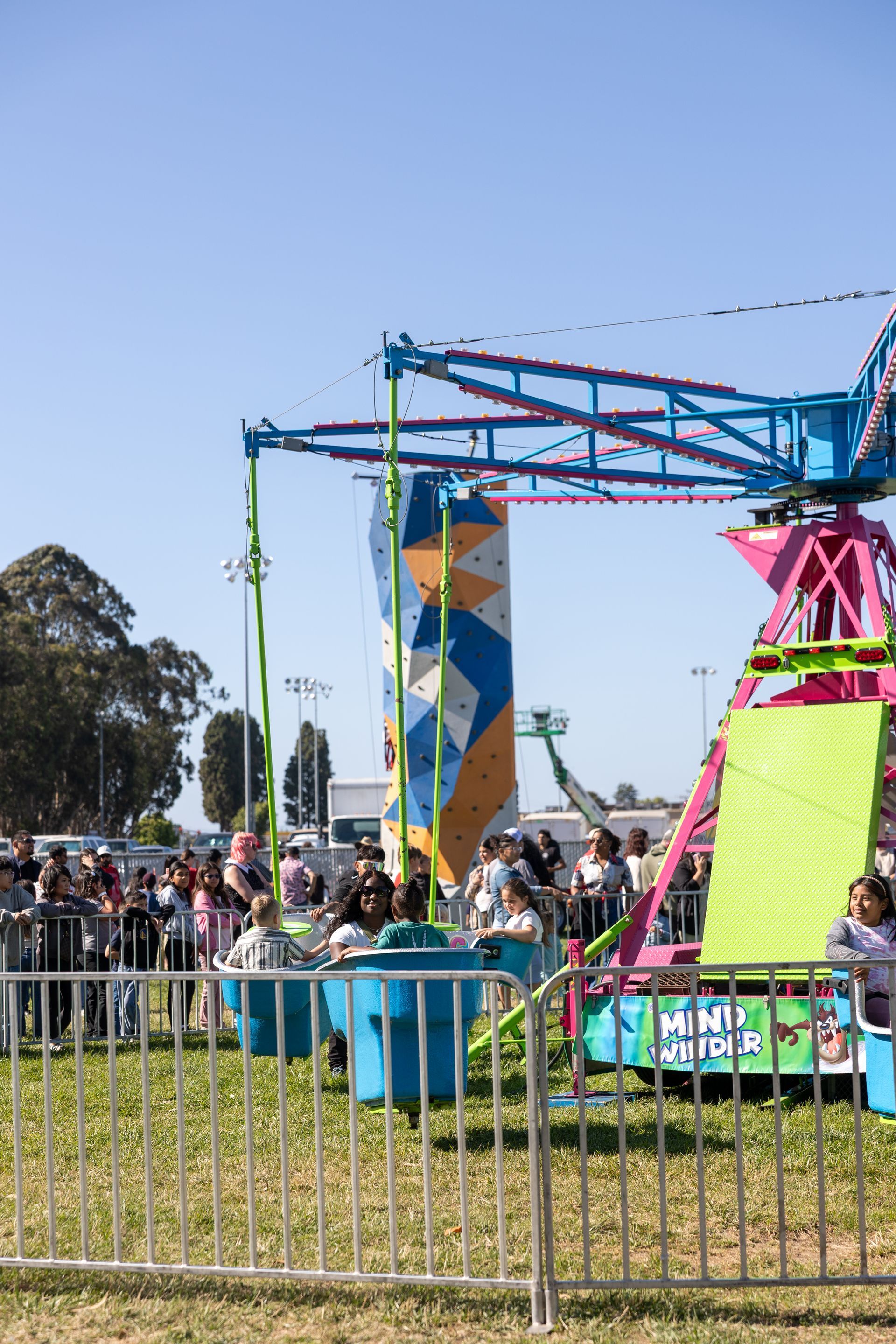 Carnival ride with people spinning on blue swings, tall climbing wall in background.