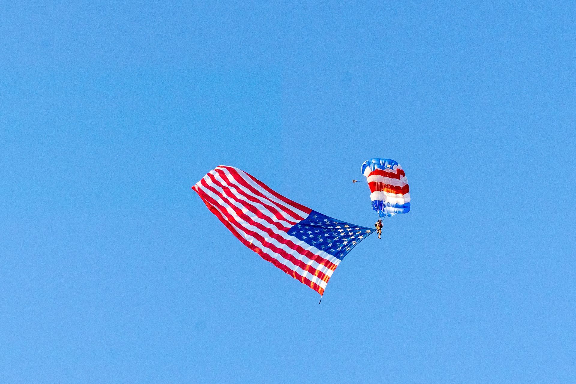 A skydiver carrying a large American flag and a red, white, and blue parachute against a blue sky.