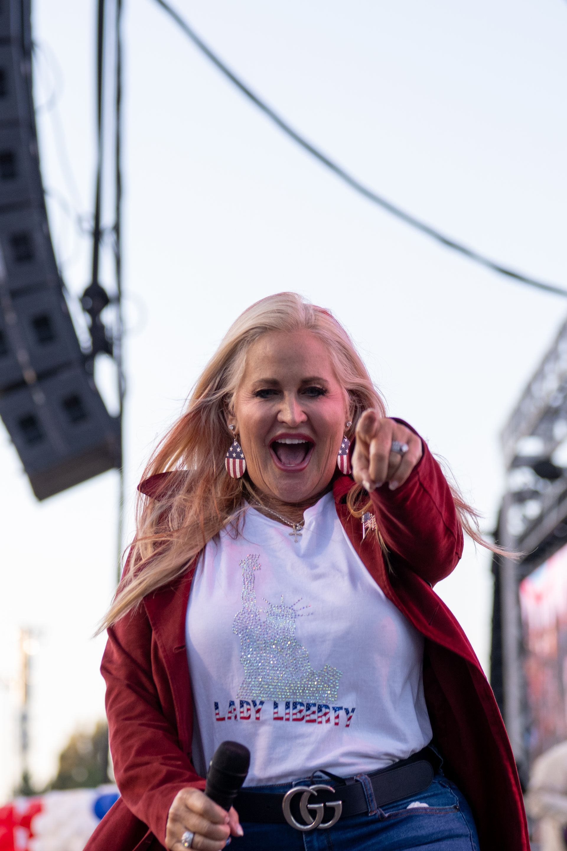 Woman with blonde hair wearing a red jacket and t-shirt pointing forward, on a stage with a microphone.