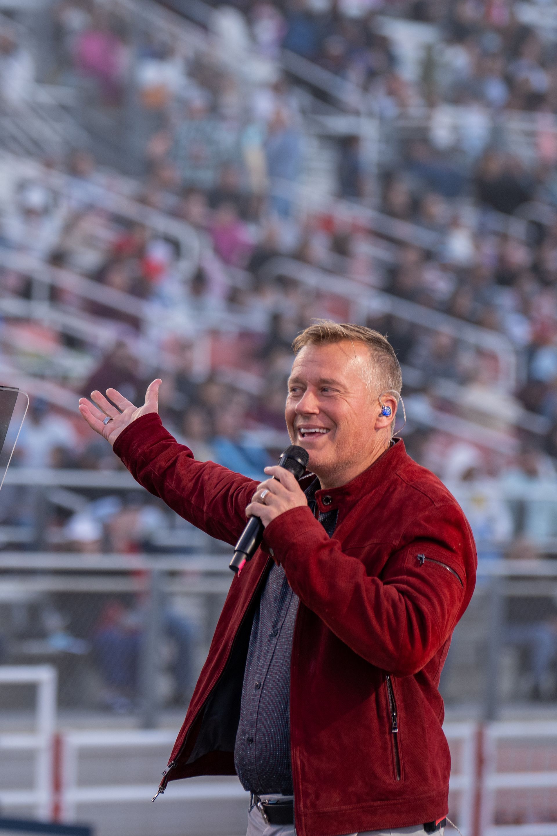Man in red jacket singing into microphone, gesturing to a crowd in a stadium.