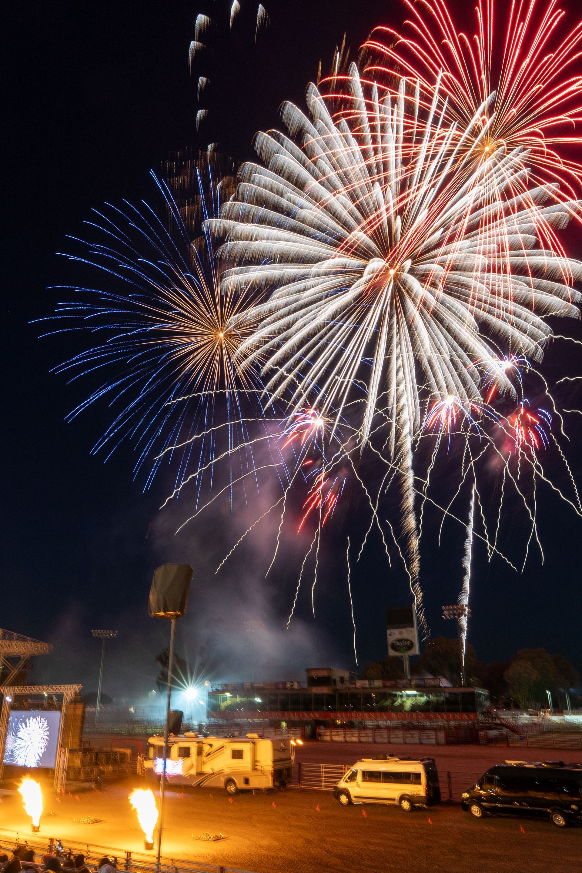 Fireworks exploding in the night sky with blue, white, and red bursts over a parking area with vehicles.