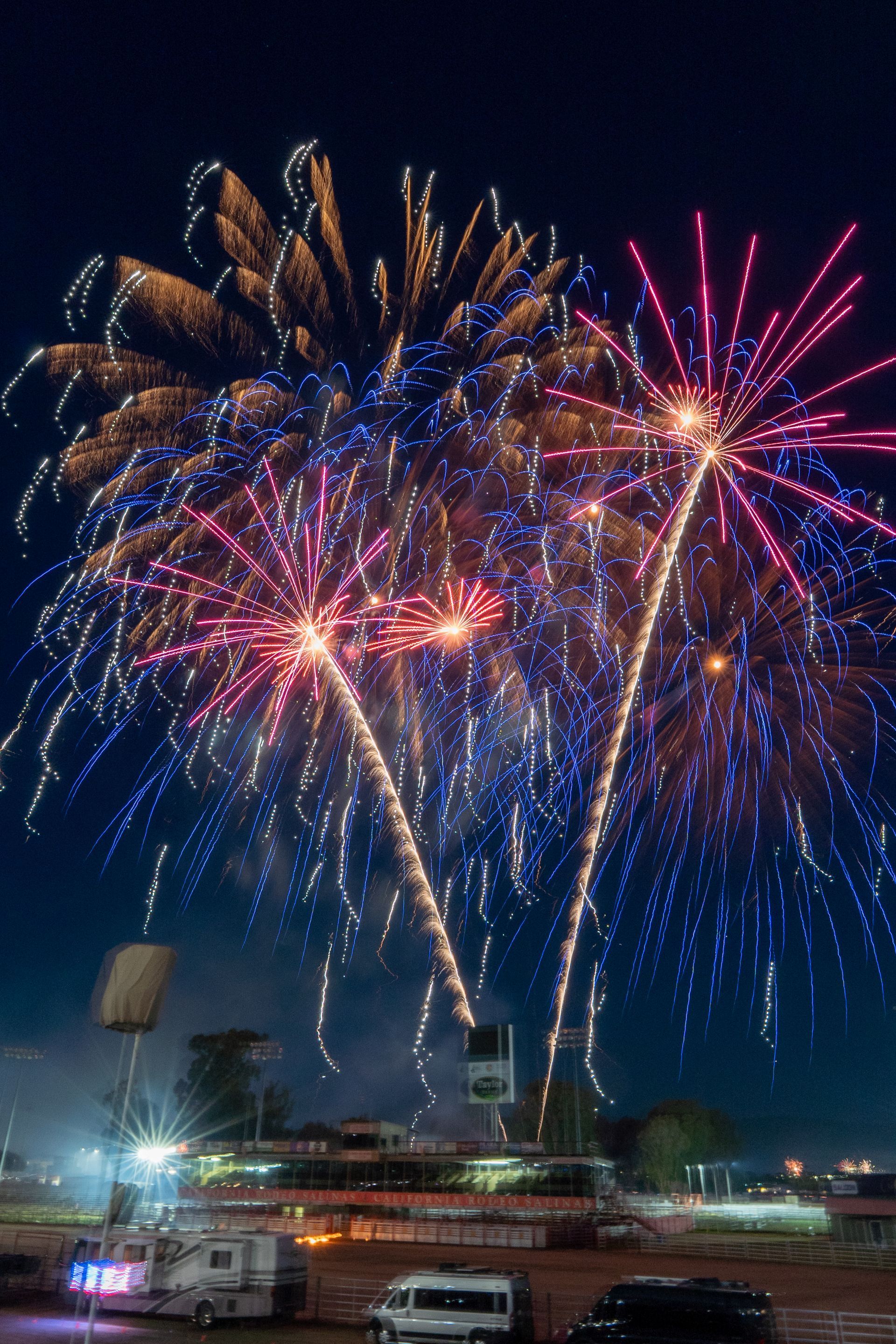 Fireworks exploding in a night sky over a fairground, displaying red, blue, and gold bursts.
