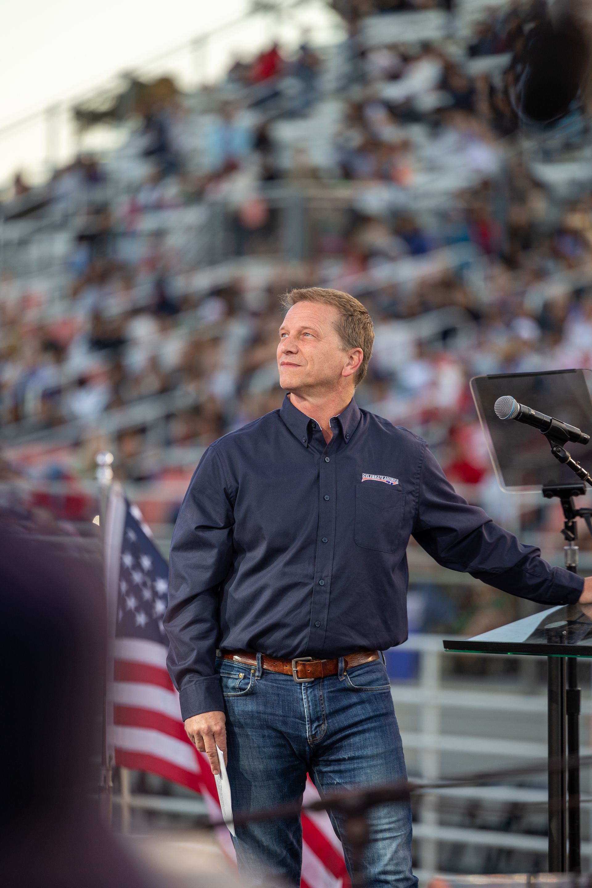 Man in blue shirt and jeans at a podium, looking upward at a crowd with flags in background.